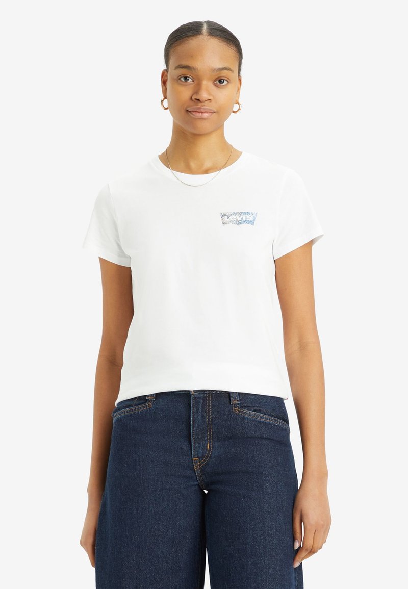T-shirt en coton blanc avec un col rond et des manches courtes, arborant un motif graphique bleu sur la zone gauche de la poitrine. Porté avec un pantalon en denim foncé.