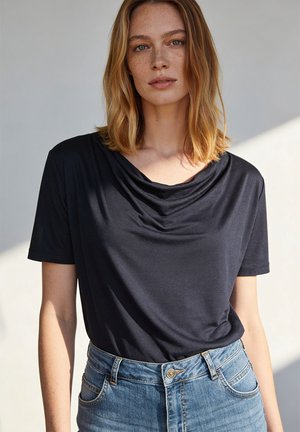 Vrouw met schouderlang blond haar die een zwart topje met korte mouwen en hooggetailleerde blauwe spijkerbroek draagt, staand tegen een lichte achtergrond.