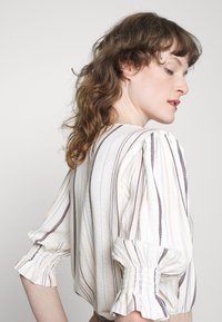 Blusa com mangas bufantes, de base branca, com riscas verticais multicoloridas e punhos franzidos. Tecido texturizado com um padrão subtil.