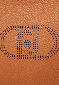 Superficie in pelle color tan con una finitura testurizzata e un design del logo perforato in nero, che mostra dettagli intricati e una consistenza liscia.