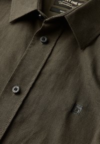Tmavě olivově zelená texturovaná košile s límečkem na knoflíky, dvěma černými knoflíky a malým čtvercovým logem na hrudi.