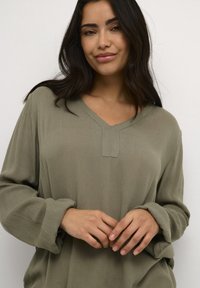 Olivgrön långärmad topp med V-hals och en texturerad yta. Har rullade manschetter och en liten rektangulär detalj ovanför ringningen.
