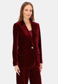 Bordowy aksamitny blezer z klapą z wycięciem, zapinany na jeden guzik, z dwoma przednimi kieszeniami. Guzik zdobiony ozdobą.