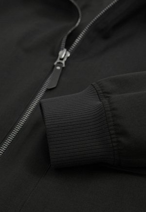 Schwarze Jacke aus glattem Material mit einem silbernen Reißverschluss und gerippten Bündchen, die ein modernes Design und eine taillierte Passform betont.