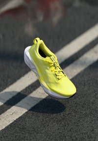 Helle gelbe Lauf shoe mit strukturiertem Obermaterial und weißer Sohle, ausgestattet mit Schnürsenkeln und einer kleinen Fersenschlaufe, schwebend über einer Gehwegoberfläche.
