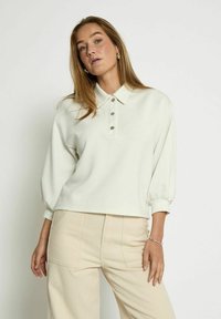 Light beige cropped pullover with a collar, three button closure, and puffed sleeves. Fabric appears soft with a smooth texture.