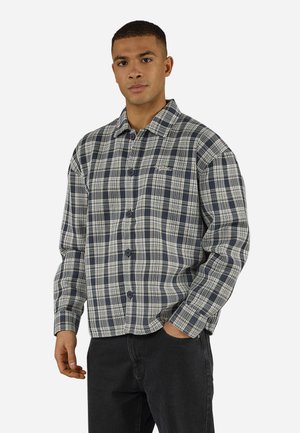 Jeune homme aux cheveux courts portant une chemise à carreaux grise et bleu marine boutonnée et un pantalon noir, debout avec une main dans la poche.