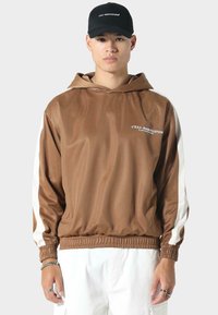 Bruine hoodie met witte zijsstrips, elastische manchetten en zoom, en een tekstgrafiek aan de voorkant. Draagt het met witte broek en een zwarte pet.