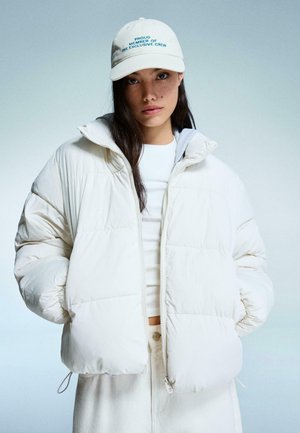 Weiß gepolsterte Jacke mit Reißverschluss, hohem Kragen und elastischen Bündchen. Getragen über einem weißen Top. Mütze mit gesticktem Text in hellbeige.