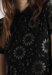 Svart spetsblus med blommönster, korta ärmar och krage. Texturen är invecklad och lägger till lager i designen.