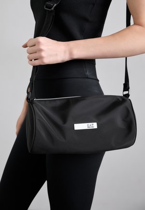 Sac en nylon noir de forme rectangulaire, fermeture éclair, bandoulière réglable et étiquette de logo blanche. Texture lisse et design minimaliste.