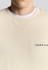 Felpa color crema con collo a girocollo a coste, dotata di un piccolo logo ricamato scuro sul lato sinistro e una texture liscia.