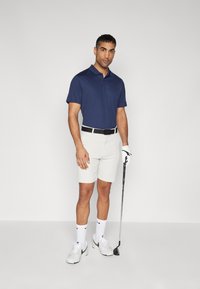 Kék polo, fehér sort, fekete öv, fehér zokni és edzőcipő. Golfütőt tart, egy sima világos háttér előtt.