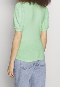 Top verde chiaro a coste con maniche a sbuffo corte, caratterizzato da un design aderente e un tessuto morbido e testurizzato. Abbinato a jeans in denim azzurro.