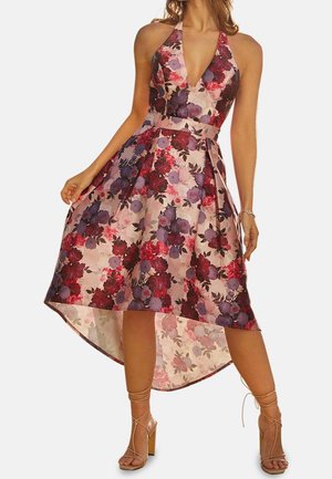 Vrouw draagt een mouwloze halterjurk met een hoog-laag zoom, voorzien van een rood, paars en roze bloemmotief, gecombineerd met sandalen met bandjes in een naaktkleur.