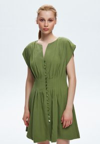 Grünes Leinenkleid mit kurzen Ärmeln, tiefem V-Ausschnitt, vertikalen Falten und Knopfverschluss vorne. Glatte Textur und knielanges Design.