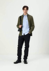 Cardigã verde tricotado, camisa azul clara de botões, calças pretas e sapatos pretos. O modelo traz uma pequena bolsa preta. Design simples, sem padrões.