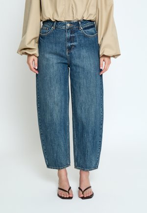 Frau trägt hoch taillierte, weit geschnittene blaue Jeans, beige Langarmbluse und schwarze Sandalen mit Absatz und steht vor einem einfarbigen Hintergrund.