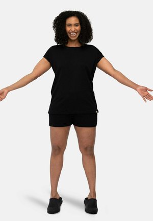 Schwarzes Kurzarmshirt mit lockerer Passform, kombiniert mit schwarzen Shorts. Das Modell trägt schwarze Slipper und steht mit ausgestreckten Armen.