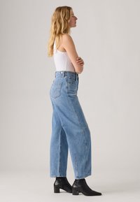 High-waisted light blue denim jeans with a relaxed fit, side slits, and back pockets paired with black heeled ankle boots.