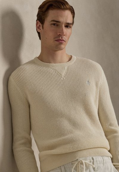 Πλεκτό πουλόβερ σε κρεμ απόχρωση με υφή, στρογγυλή λαιμόκοψη και ριμπ μανσέτες, φορεμένο με ανοιχτόχρωμο παντελόνι.