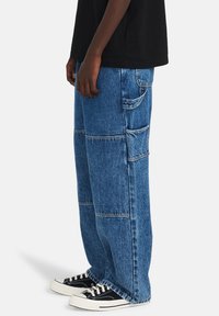 Pantalones cargo de mezclilla azul con un ajuste holgado, múltiples bolsillos, costuras en contraste y una cinturilla plana. Combinados con zapatillas negras.