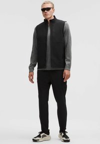 Melnā bezpiedurkņu veste virs pelēkas garas piedurknes krekla, saskaņota ar melniem uzliekamiem biksēm un melnām/kremīgām sporta apaviem. Modelis valkā saulesbrilles.