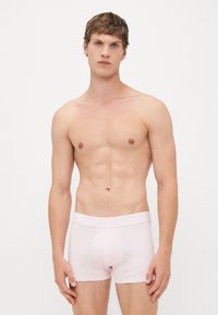 Jonge man met kort bruin haar, gekleed in lichtroze Lacoste onderbroek, staand tegen een plain witte achtergrond.