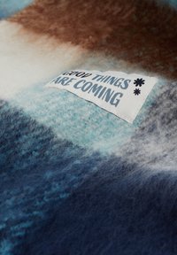 Volnena odeja z gradientom modrih, rjavih in smetanastih odtenkov, z všitom etiketom, ki pravi "DOBRE STVARI PRIHajajo" v modri barvi.