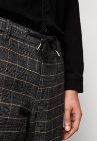 Karierte graue und schwarze Hose mit Kordelzugbund und Metallenden. Der Stoff hat eine strukturierte Oberfläche und ein sichtbares Webmuster.