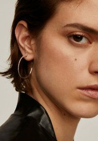 Pendiente de aro plateado con un borde texturizado que presenta pequeñas piedras incrustadas, llevado por una persona con cabello corto y oscuro que viste una chaqueta de cuero negra.