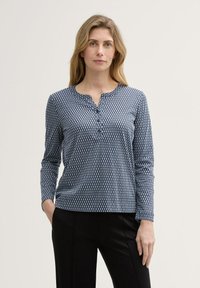 Marineblå langærmet skjorte med et geometrisk hvidt mønster, med en henley-udskæring og blødt, struktureret stof. Sammen med sorte bukser.