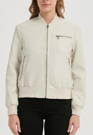 Femme portant une veste bomber crème zippée avec des poches zippées et des poignets côtelés, associée à un pantalon noir.
