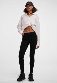 Jeans slim-fit noirs en denim, associés à une chemise blanche nouée et une casquette noire. Baskets noires à semelles épaisses.