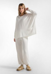 Witte gebreide oversized trui met een ronde halslijn, voorzien van een zak aan de voorkant, gecombineerd met bijpassende witte broek en tan slippers.