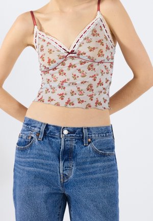 Persona con un top camisola floral translúcido con detalles de encaje y vaqueros azules de cintura alta, manos apoyadas detrás de la espalda.
