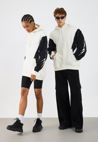 Twee modellen dragen oversized hoodies met een crème kleurige body en zwarte mouwen met een grafisch ontwerp. De outfit wordt aangevuld met zwarte shorts en broeken.