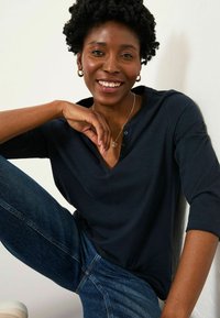 Navy blue, cotton blend top with a V-neck and three-quarter sleeves. Model wears light blue denim jeans, seated against a white wall.