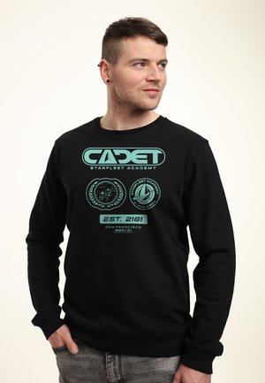 Man draagt een zwarte trui met turquoise "CADET Starfleet Academy" tekst en logo's, staat met één hand in de zak, kijkt naar rechts.