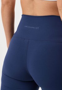 Primer plano de una persona que lleva leggings deportivos azul marino de talle alto con el logo de "Swedemount" en la banda trasera de la cintura.