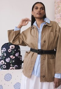 Barna pamut dzseki nagy zsebekkel, fekete övvel megkötve; világoskék ing alatta. Virágmintás sötétkék hátizsák mellett.