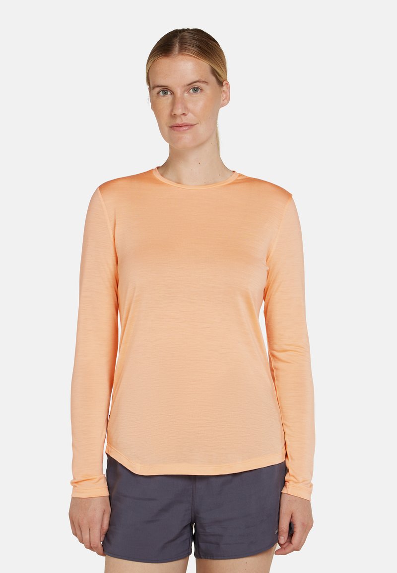 Kvinna i en persikofärgad långärmad skjorta och mörkgrå shorts, stående mot en enfärgad vit bakgrund.