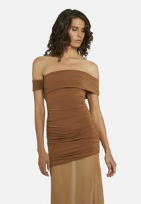 Vrouw in een bruin off-the-shoulder gerimpeld jurk met doorzichtig materiaal aan de onderkant, staand met ontspannen armen en naar beneden kijkend.