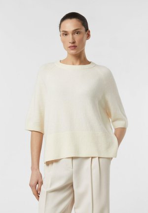 Frau mit dunklen, zurückgekämmten Haaren, die einen cremefarbenen Kurzarm-Pullover und passende plissierte Hosen trägt und mit einer Hand in der Tasche steht.