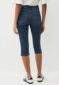 Tummansiniset farkku capri-housut, joissa on kaksi takataskua, käännetyt helmat ja tiukka malli, yhdistettynä valkoisiin lenkkareihin, joissa on mustia yksityiskohtia.