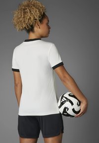 White ribbed football shirt with a black collar and cuffs, paired with black shorts. A black-and-white soccer ball is held against the body.