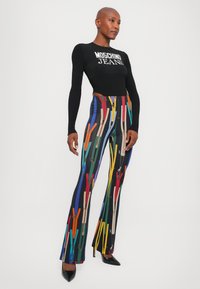 Čierny dlhý rukáv top s bielym nápisom "Moschino Jeans"; farebné vzorované rozšírené nohavice s abstraktnými strihovými vzormi; vysoké podpätky.