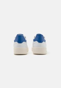 White sneakers featuring a blue heel tab, smooth leather material, rounded shape, and textured rubber sole with a light beige finish.