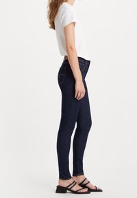 Jeans ajustados de color azul oscuro, material de denim, de tiro alto, con bolsillos laterales y traseros con detalle de costura, combinados con una camiseta blanca y sandalias negras.