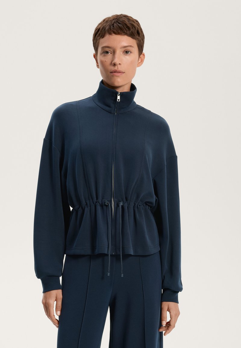 Mornarica zip-up jakna s visokim ovratnikom, skupljenim strukom i dugim rukavima, izrađena od glatke tkanine. Sadrži detalje s vezicama i zip zatvaranje.